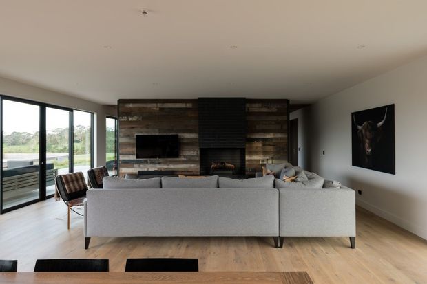 Open-plan living room viewed from behind a large light-grey sectional sofa, with a rustic reclaimed-timber feature wall, dark tiled fireplace surround and built-in television. Full-height black-framed glazing lines one side of the room, while pale timber flooring and restrained contemporary furnishings create a spacious, warm interior.