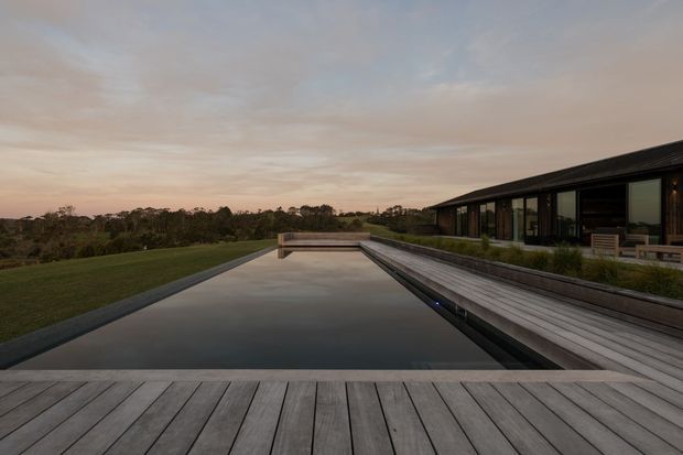 A contemporary single-storey residence sits beside a long, narrow reflecting pool or swimming pool. Wide weathered timber decking frames the pool, while the dark linear home has extensive floor-to-ceiling glazing and a low-pitched roof. The landscaped lawn and distant tree line are viewed at dusk under a softly clouded sky.