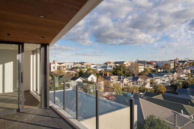 Elevated balcony or terrace looking across a dense suburban neighbourhood toward a distant body of water. The foreground features frameless glass balustrades with dark metal posts, large sliding or folding glazed doors, dark tiled flooring, and a warm timber-lined soffit beneath a broad roof overhang. The view suggests a modern multi-level residence with expansive outdoor living and a wide urban outlook.