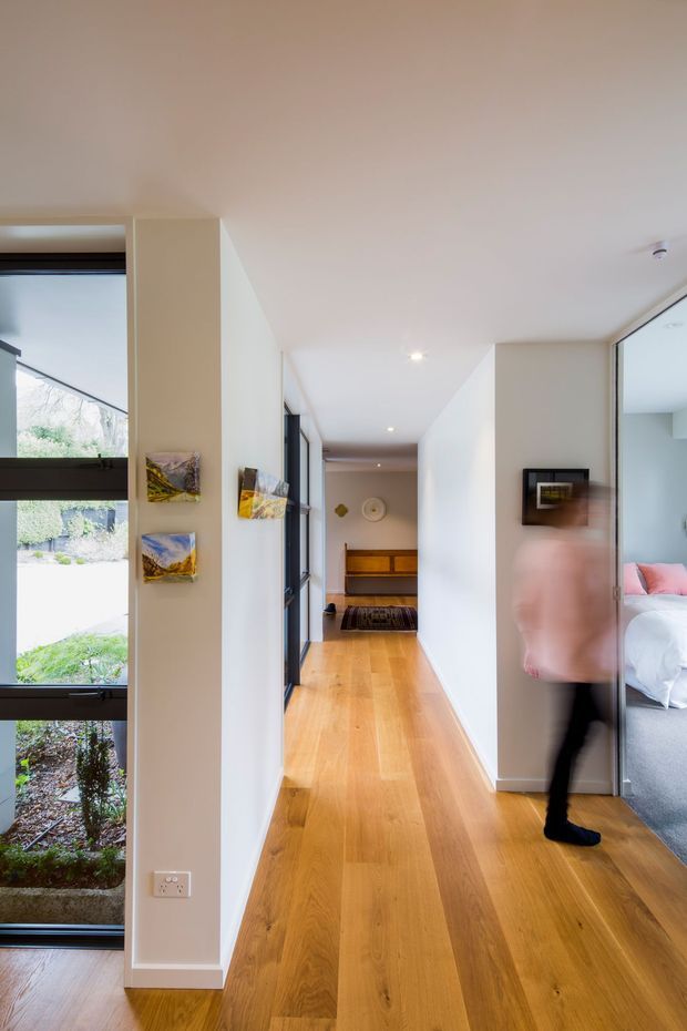 A bright contemporary residential hallway with warm timber flooring, white walls and ceiling, recessed downlights, and extensive dark-framed glazing. The corridor leads toward a timber console and bench, with a bedroom visible through a glazed opening on the right. Small artworks decorate the central wall, and a blurred person is walking through the space.