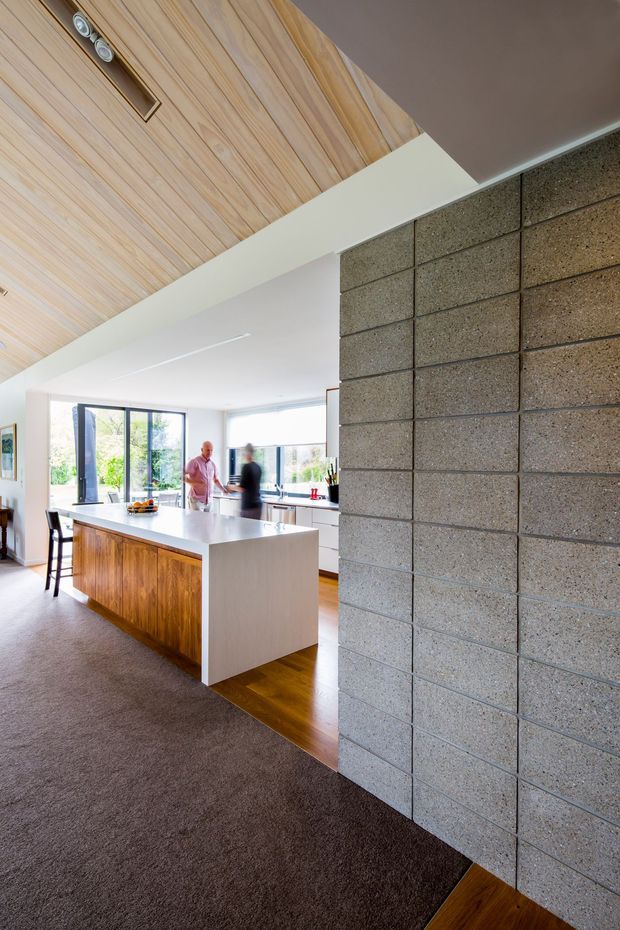 A bright open-plan kitchen viewed from an adjoining living area, featuring a large white island with warm timber cabinetry, expansive black-framed glazing, a timber-lined angled ceiling with recessed spotlights, and a prominent exposed grey concrete block wall. Dark carpet transitions to polished-looking timber flooring in the kitchen.