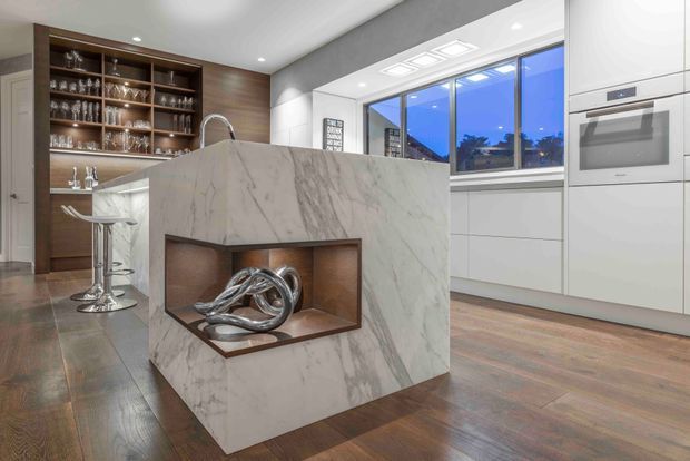 Contemporary kitchen featuring a large veined stone island with an integrated display niche, white built-in cabinetry and appliances, dark timber flooring, and a timber bar area with open glassware shelving. Floor-to-ceiling windows and recessed ceiling lighting create a bright, refined interior.