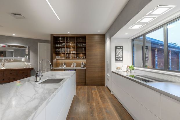 Contemporary kitchen with a large white marble-look island, integrated sink and tapware, extensive white cabinetry, timber flooring, and a built-in timber drinks cabinet with open shelving. A wide window and streamlined linear lighting create a bright, refined space.