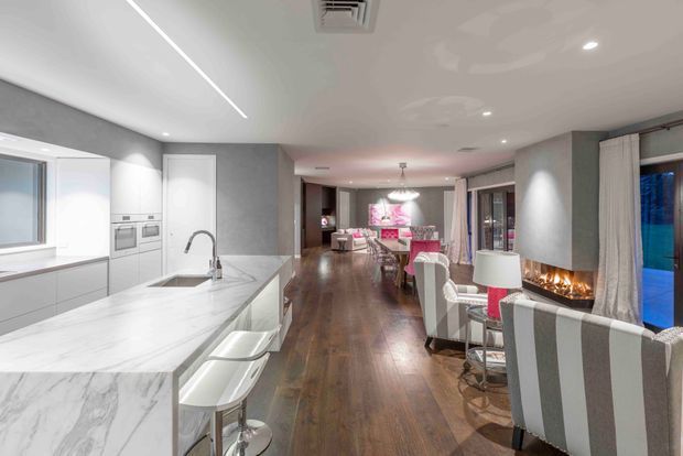 Open-plan contemporary interior combining a white marble-look kitchen with island seating, a dining area, and a living zone. The space features dark timber flooring, pale grey walls, white cabinetry, recessed lighting, floor-to-ceiling curtains, and a linear fireplace set into a grey feature wall. Large glazing at the right provides blue-toned exterior views.
