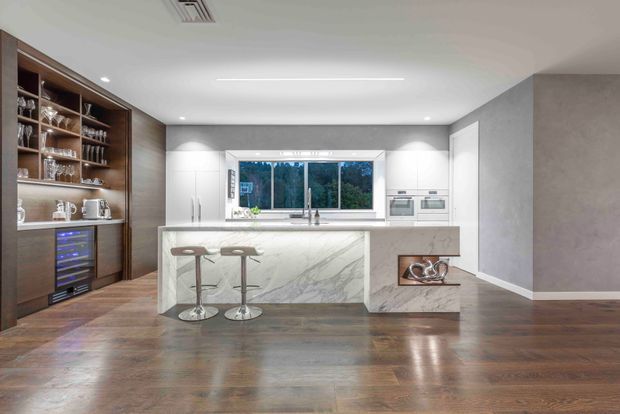 A refined contemporary kitchen featuring a large marble-look island with integrated seating, white cabinetry, timber open shelving and a built-in beverage or wine fridge. A window above the sink provides natural light and views of greenery, while warm timber flooring contrasts with the predominantly white and grey fit-out.