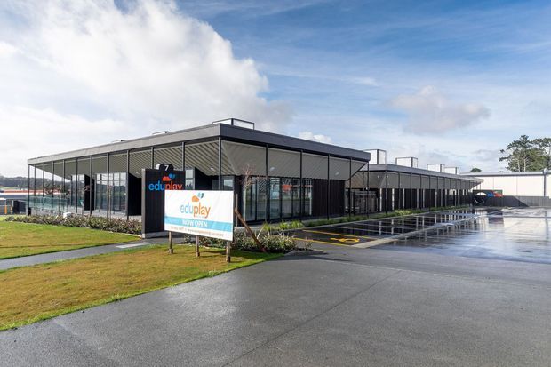 Exterior view of a contemporary single-storey commercial building with a long, low flat-roof form, dark metal cladding, extensive glazed frontage, covered entry, landscaped lawn, and wet paved parking/drop-off areas. Signage indicates an education and childcare/play facility.