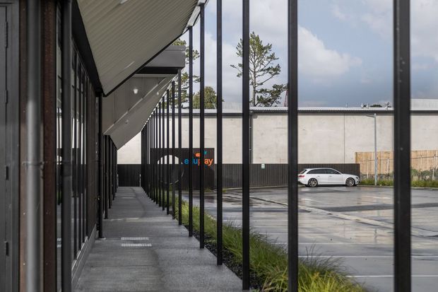 A contemporary commercial or industrial building frontage viewed along a covered pedestrian walkway. Slim black steel posts and full-height glazing line the left side, with angled roof overhangs above and a paved parking area visible beyond a low landscaped strip. The scene uses restrained industrial materials and a neutral grey palette.