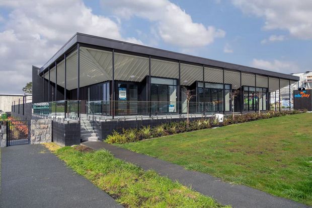 A single-storey contemporary commercial building with a broad low-pitched roof, dark structural framing, extensive glazing, and a covered entrance veranda. The building sits beside paved pedestrian paths and landscaped lawn, with signage suggesting an education or childcare facility.