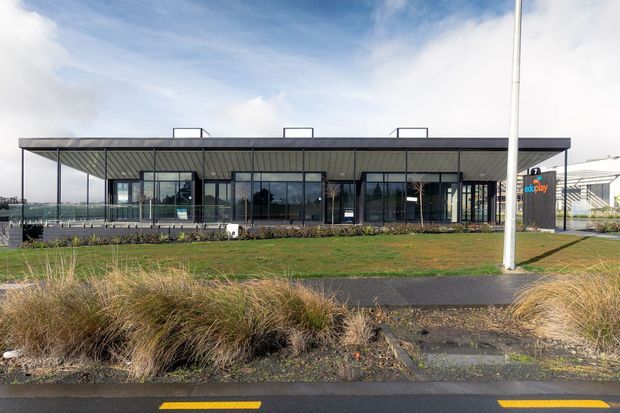 Single-storey contemporary commercial building viewed from across a landscaped roadside area. The building has a long, low rectangular form with extensive floor-to-ceiling glazing, dark metal framing, a broad flat roof with a deep projecting canopy, and rooftop mechanical elements. A sign and covered structure are visible at the right, with native-style grasses and paved paths in the foreground.