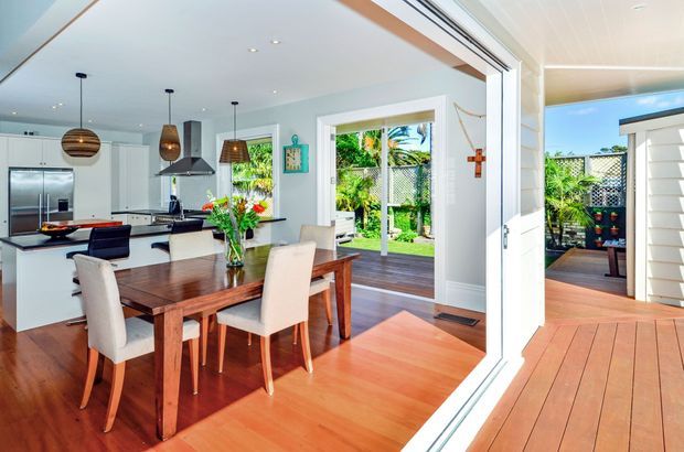 Bright open-plan dining and kitchen interior opening onto a timber deck and landscaped garden. The room features warm timber flooring, a dark timber dining table with upholstered chairs, white cabinetry, stainless-steel appliances, pendant lighting, and large glazed openings creating a strong indoor-outdoor connection.