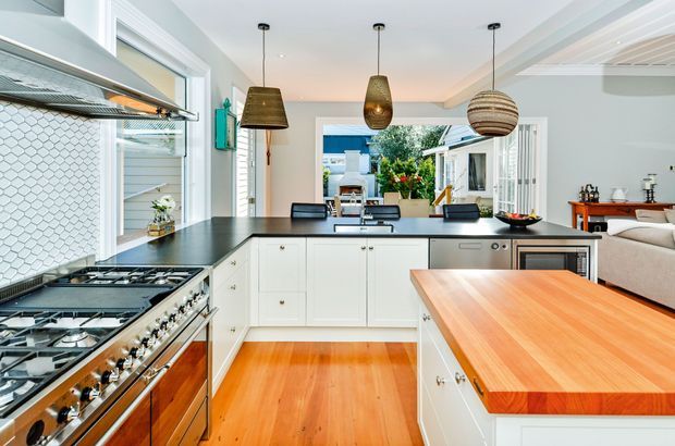 Bright, spacious contemporary kitchen with white shaker-style cabinetry, a dark stone island benchtop, warm timber flooring, and a large timber-topped peninsula. A professional-style stainless steel range and sculptural pendant lights are prominent features. The kitchen opens toward a dining and living area and a glazed outdoor space beyond.