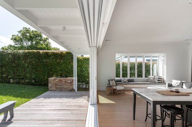 Bright open-plan interior opening directly onto a timber deck and landscaped backyard through fully retractable glazed doors. The room has white walls and ceiling, pale timber flooring, a large dining table with stools, and a built-in window seat with cushions. The covered outdoor area includes a brick planter or low wall and a neatly clipped hedge.
