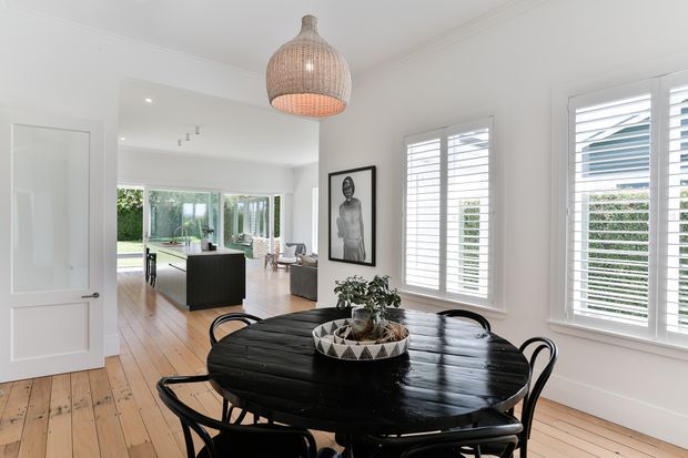 Bright dining room with a large round black timber dining table and black chairs, white walls and plantation shutters, wide timber floorboards, a woven pendant light, and a framed artwork. An open doorway leads toward a kitchen and glazed living area beyond.