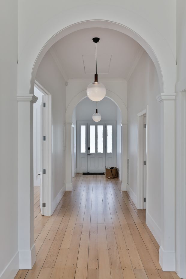 A bright, refined residential hallway with a long perspective toward a glazed front door. The corridor features white painted walls, ornate arched openings with classical moulded trim, repeated pendant globe lights, and wide pale timber floorboards. Open doorways on both sides lead to adjacent rooms.