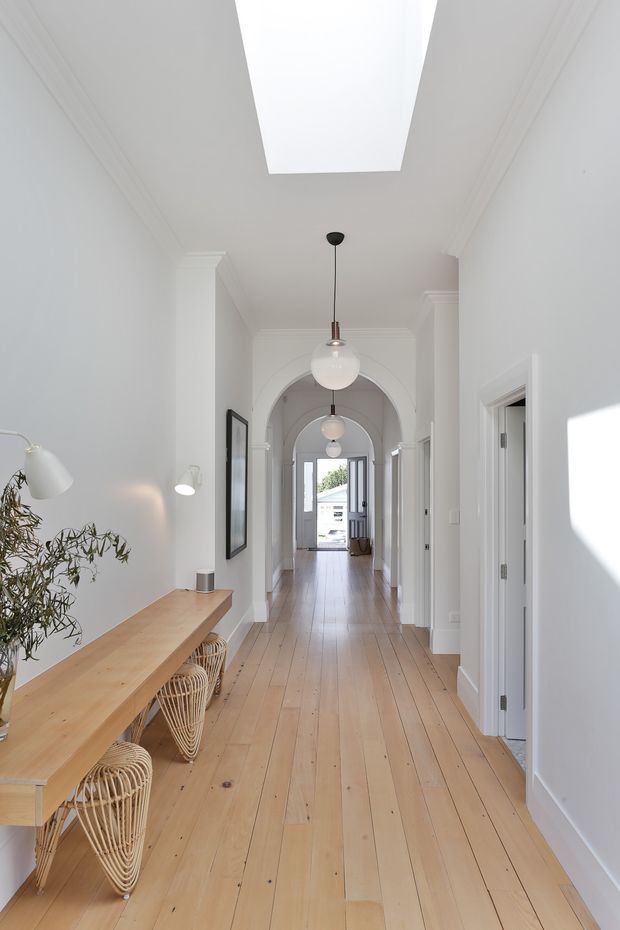 Bright, elongated residential hallway with white walls, decorative ceiling mouldings, repeated arched openings, and a large skylight. Wide pale timber floorboards lead toward a glazed entry at the far end. A light timber console with woven rattan stools occupies the left side, with framed artwork, pendant globe lights, and a potted plant adding detail.