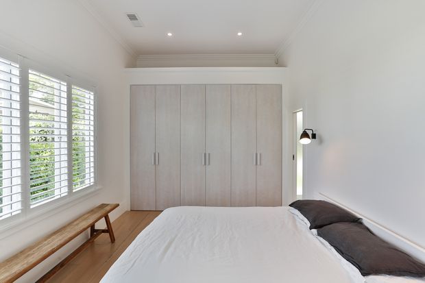 Bright, serene bedroom with a large built-in pale timber wardrobe, white bedding, a dark accent cushion, timber flooring, plantation shutters, and classic decorative ceiling cornices. A simple timber bench sits beneath the windows, with a wall-mounted bedside light.