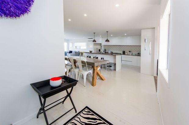 Bright open-plan interior showing a contemporary kitchen and dining area. White cabinetry, pale flooring, a tiled splashback, recessed ceiling lights, pendant lights, a timber dining table, and metal dining chairs create a clean, airy space. A living area is visible beyond the dining zone, with a console table and patterned rug in the foreground.