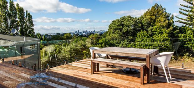 An elevated outdoor timber deck with a built-in glass safety balustrade, furnished with a large wooden dining table and bench seating. The deck overlooks a leafy suburban landscape and distant city skyline under a bright blue sky.