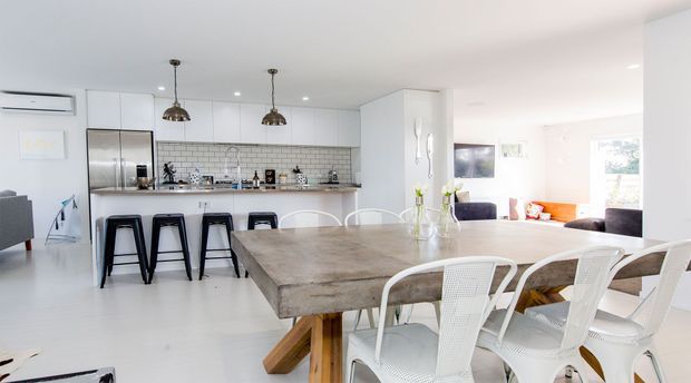 Bright open-plan kitchen and dining area with a large rustic timber dining table, white woven dining chairs, a white kitchen with stainless-steel appliances, tiled splashback, breakfast bar and pendant lights. A living area is visible beyond the dining space.