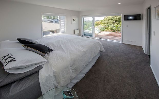 A bright, contemporary bedroom with a large bed dressed in white bedding and dark accent pillows, grey carpet, white walls, recessed ceiling lights, and a wall-mounted television. Wide sliding glass doors open to a timber outdoor deck, with additional glazing and shutters providing natural light.