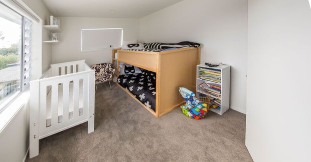 Bright children's bedroom with a white cot, wooden bunk bed, patterned bedding, shelving, toys, carpeted floor and large windows.