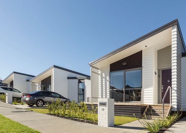 Street-level exterior view of a row of contemporary single-storey homes with white horizontal cladding, dark pitched roof edges, recessed entry porches, large windows, concrete driveways, landscaped front gardens, and prominent white brick letterboxes. The foreground dwelling has a dark timber-look entry wall and a visible house number 19.