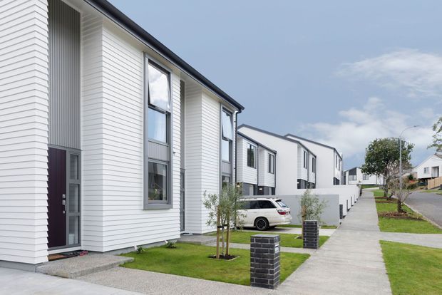 A streetscape view of a contemporary residential development with a row of narrow multi-storey homes. The houses feature white horizontal cladding, dark window and roof trims, tall vertical glazing, recessed entry doors, small front lawns, paved walkways, and low dark-brick entry pillars. The development sits on a gently sloping suburban street beneath a partly cloudy sky.