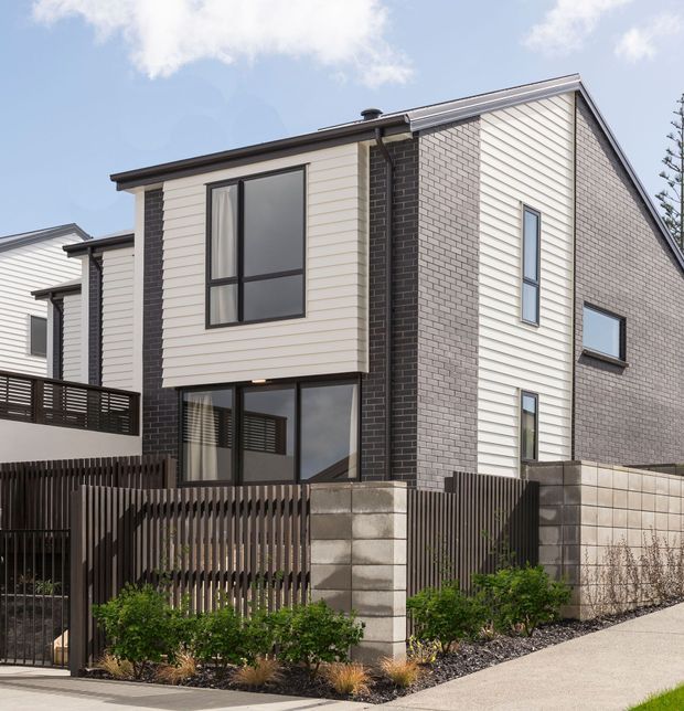 Exterior view of a contemporary two-storey townhouse with a simple pitched roof, contrasting light horizontal cladding and pale brickwork, dark brick vertical feature panel, black-framed glazing, and a small landscaped front courtyard enclosed by vertical timber fencing and concrete block walls.