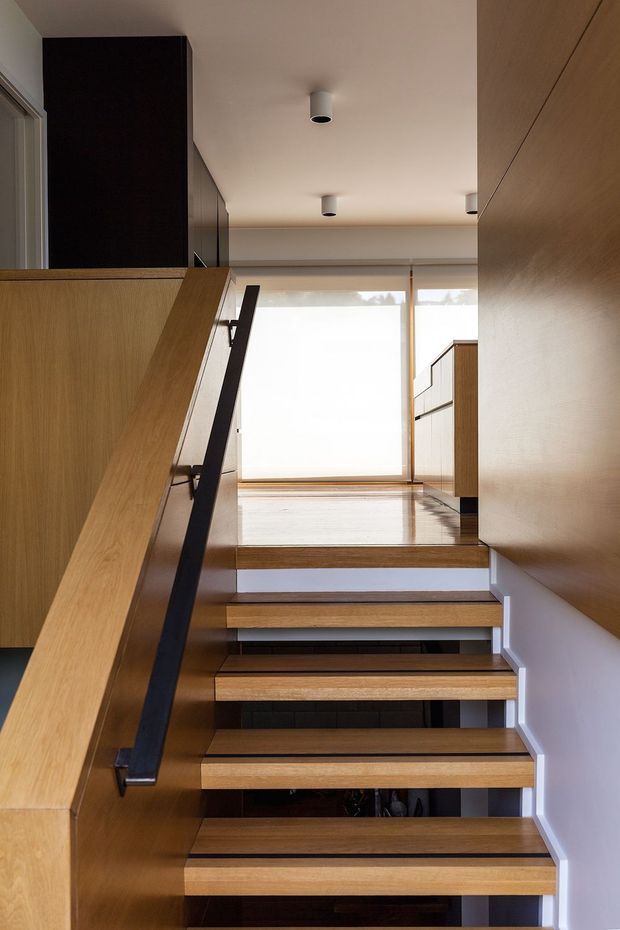 A modern interior stairway viewed from the lower level, with open-riser timber steps, a simple black metal handrail, and warm timber wall and balustrade surfaces. The stairs lead to a bright landing with large translucent glazing, while recessed cylindrical ceiling lights and clean white walls create a minimalist, contemporary atmosphere.