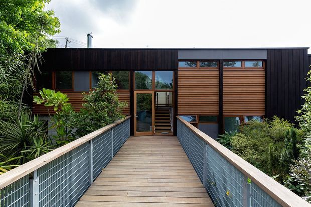 Exterior view of a contemporary single-storey building approached by a timber pedestrian bridge. The facade combines dark vertical cladding, warm horizontal timber screening, and large timber-framed windows, surrounded by dense landscaped planting.