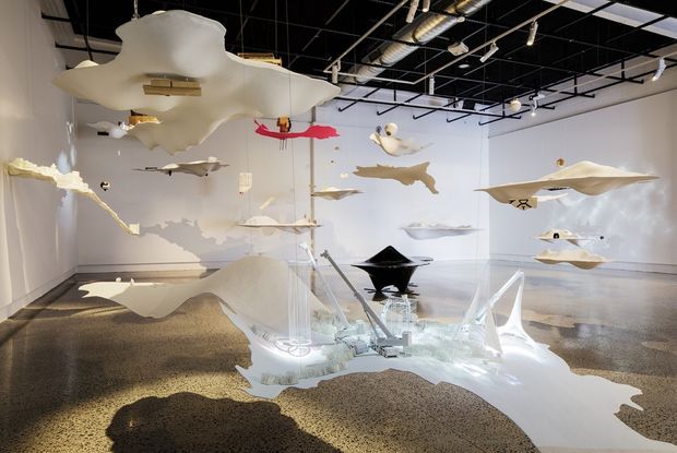 A spacious contemporary gallery or exhibition interior with white walls, polished concrete flooring, exposed black ceiling services, and numerous suspended sculptural or installation pieces floating throughout the room. The artwork casts dramatic shadows across the floor, creating an immersive and highly artistic display.
