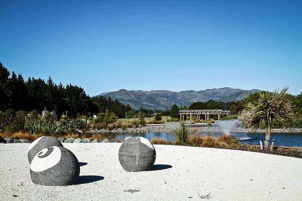 A landscaped outdoor garden and water feature beside a broad white gravel area, with two large dark spherical sculptures in the foreground. Ornamental grasses, palm-like plants, trees, a pergola, and a lake or pond sit against a mountain backdrop under a clear blue sky.