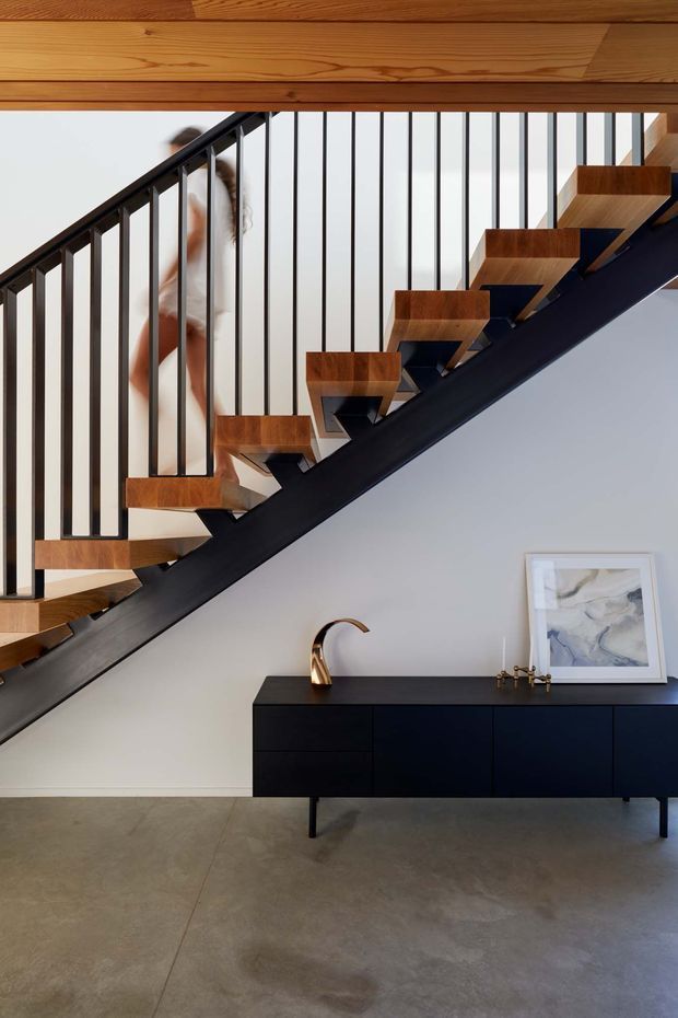 Interior view of a contemporary stairway with floating timber treads, a dark steel stringer, and slim black vertical balustrade rods. A timber-lined ceiling is visible above. Beneath the stairs is a minimalist black sideboard with a sculptural brass tap or vessel, small decorative objects, and framed artwork. The room has pale walls and a polished grey concrete floor.