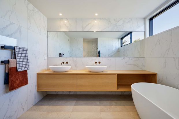 Bright contemporary bathroom with a floating double vanity in light timber, two white vessel basins, a large wall-to-wall mirror, marble-look wall surfaces, warm stone-look floor tiles, and a freestanding white bathtub. High-set glazing brings in natural light.