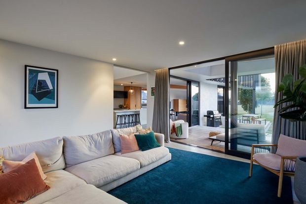 Contemporary open-plan living room with a large light sectional sofa, deep teal carpet, and colourful cushions. Full-height glazed sliding doors open the room to an outdoor terrace, while a compact kitchen and dining area are visible beyond. Neutral walls, recessed ceiling lights, full-height curtains, and abundant indoor greenery create a relaxed, polished interior.