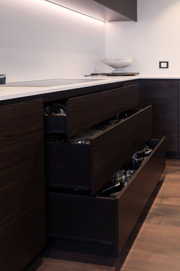 Close-up view of a refined kitchen featuring extensive dark wood-grain drawer cabinetry with multiple drawers open to reveal organised cookware and utensils. A pale stone benchtop runs along the cabinetry, paired with a smooth light wall and discreet linear under-cabinet lighting. The space has warm timber flooring and minimal decorative styling, including a shallow bowl on the benchtop.