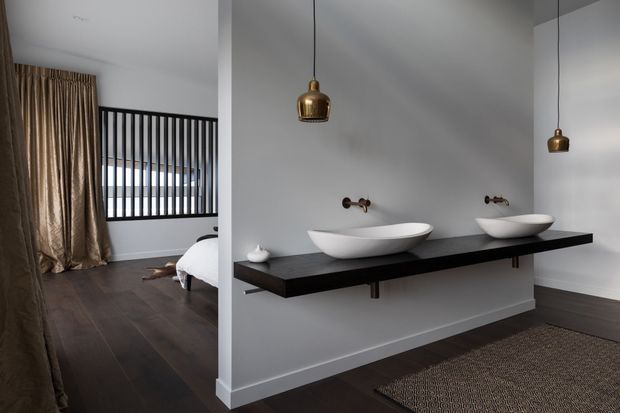 Contemporary bathroom vanity with two white vessel basins on a long dark floating benchtop, wall-mounted bronze tapware, and pendant lights. The bathroom is open to a bedroom visible on the left, with dark timber flooring, neutral walls, full-height curtains, and a black vertical-slat balustrade.