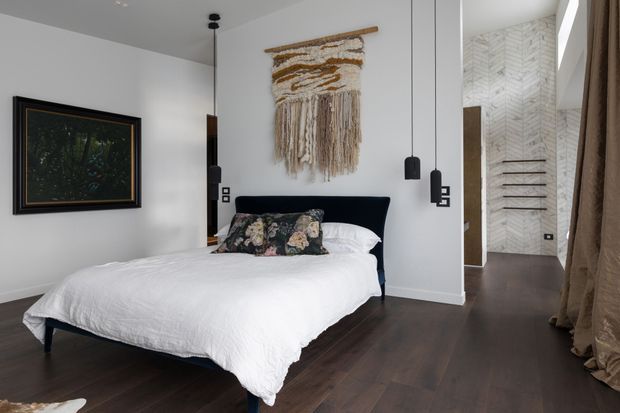 A stylish bedroom with a dark upholstered bed, white bedding, patterned decorative cushions, dark timber flooring, and white walls. A woven wall hanging serves as the main feature above the bed, complemented by framed artwork, slim black pendant lights, and full-length neutral curtains. An open passage reveals a bright tiled bathroom or dressing area with a chevron-patterned wall finish.