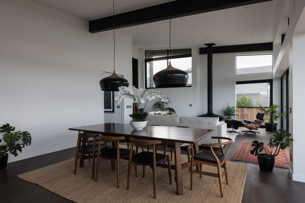 Open-plan contemporary dining and living area with a timber dining table and chairs in the foreground, a pale upholstered sofa beyond, large black-framed windows, dark timber flooring, and large black pendant lights. Indoor plants, a woven rug, and restrained furnishings create a warm, refined interior. Exposed black structural beams and generous glazing add architectural definition.