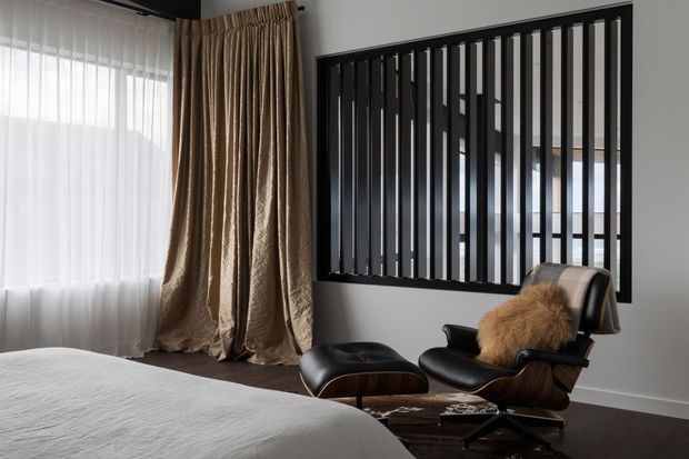 A refined contemporary bedroom with a partially visible upholstered bed, sheer white curtains layered with heavy taupe drapes, and a black leather lounge chair with matching ottoman. A large black vertical-slat interior screen or partition forms a strong architectural feature on the wall, with dark flooring and neutral walls creating a sophisticated, moody palette.