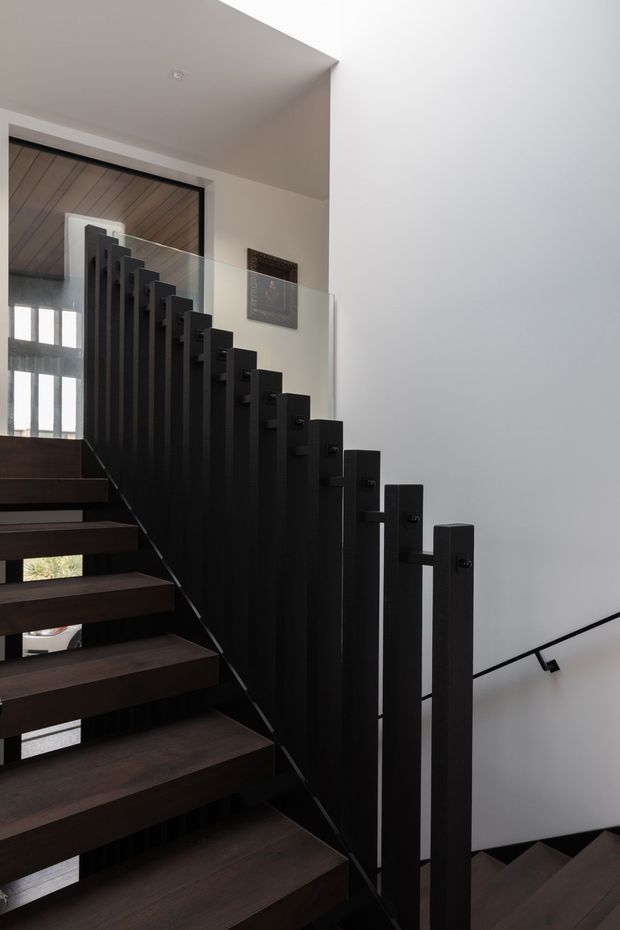 Interior view of a modern stairway with dark timber open-riser treads and a striking black vertical-picket balustrade. A clear glass panel follows the upper stair edge, while a minimalist black handrail runs along the white wall descending to the lower level. The stairwell is bright and contemporary, with a timber-lined ceiling visible at the landing.