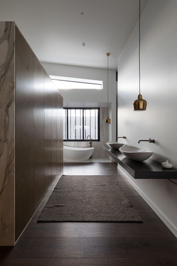 A refined contemporary bathroom viewed along a narrow vanity corridor toward a freestanding bathtub. The room features dark timber flooring, tall dark wood cabinetry on the left, a dark floating double vanity with two vessel basins on the right, white walls, a marble-look wall panel, black-framed window glazing, and warm brass pendant lights. The space is elongated, calm, and softly daylit.