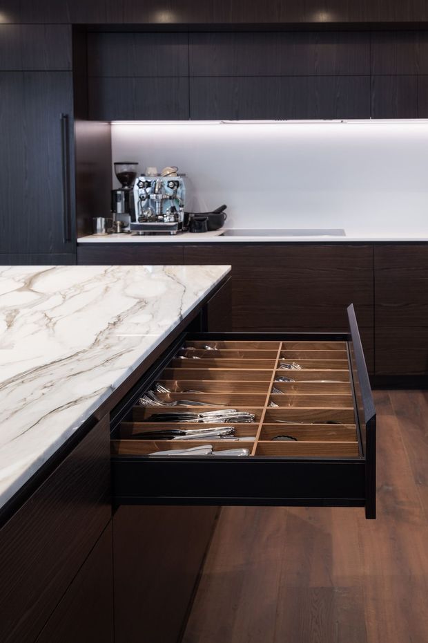 Close-up view of a dark, sophisticated kitchen featuring extensive dark timber cabinetry, an open wide cutlery drawer with timber dividers, and a pale veined stone island benchtop. A coffee machine and cooktop area sit against a softly illuminated light splashback, with timber flooring completing the space.