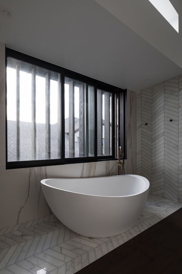 A contemporary bathroom featuring a sculptural freestanding white bathtub beneath large black-framed windows. The walls and floor are clad in white marble-look herringbone tile, with a matching marble-look wall surface beneath the windows. A slim skylight or clerestory opening brings additional daylight into the room, while dark timber flooring borders the tiled bathing area. Brushed brass or bronze plumbing fixtures add a subtle accent.