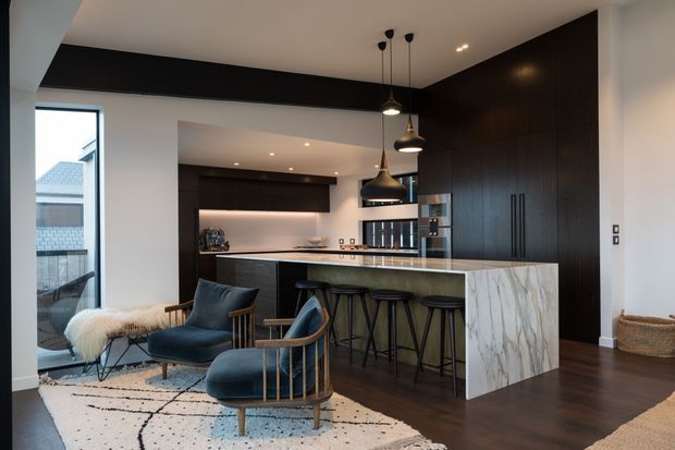Open-plan contemporary kitchen and living area with dark timber cabinetry, a large marble-look waterfall island, pendant lighting, and seating for four. The foreground includes two upholstered lounge chairs on a patterned rug, while large glazing brings in daylight and views of neighbouring buildings.