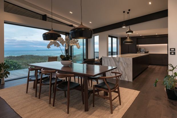 A refined open-plan dining and kitchen interior with expansive floor-to-ceiling glazing overlooking the ocean and coastal landscape. The dining area features a large dark timber table, sculptural timber dining chairs, woven rug, and statement black pendant lights. Behind it is a contemporary kitchen with dark cabinetry, a dramatic white marble island, integrated appliances, and recessed lighting. The palette is warm and sophisticated, combining dark timber, black accents, pale stone, and natural textures.