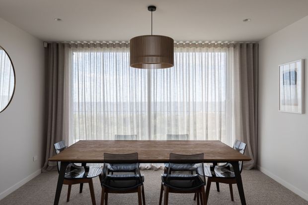 A refined dining room with a long dark timber dining table, six dining chairs, and a large cylindrical pendant light. Full-height sheer curtains and taupe drapes span the rear glazing, with neutral painted walls, carpet flooring, and framed artwork. The room has a calm, contemporary feel with soft natural light.