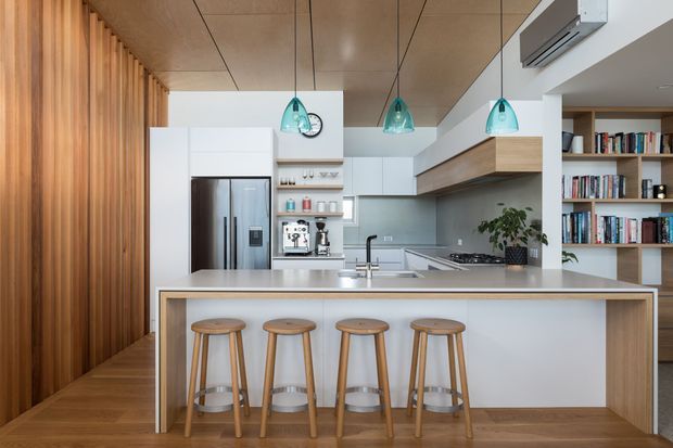 Contemporary open-plan kitchen with a large island and four timber bar stools, white cabinetry, integrated stainless-steel appliances, pale benchtops, and a grey-green tiled splashback. Warm timber wall panelling, timber flooring, and a timber ceiling add natural warmth, while turquoise pendant lights provide colour accents. A built-in bookshelf and living area are visible beyond the kitchen.