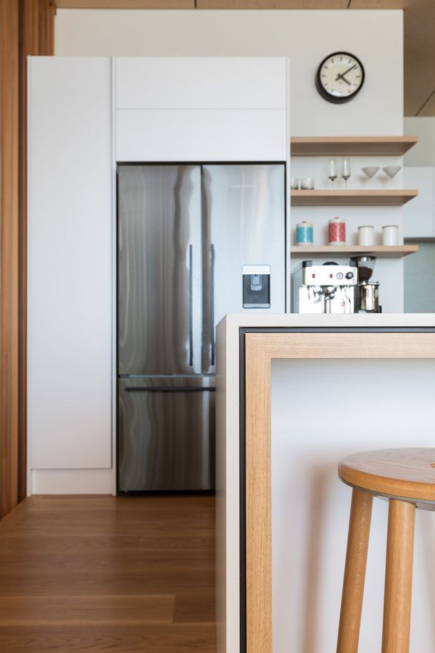 A compact contemporary kitchen featuring a tall stainless-steel refrigerator integrated into white cabinetry, open timber shelves displaying glassware and crockery, and a coffee machine. A white island or breakfast bar with light timber trim occupies the foreground, accompanied by a timber stool. Warm timber flooring and vertical timber wall detailing add natural warmth, while a round wall clock provides a small decorative accent.