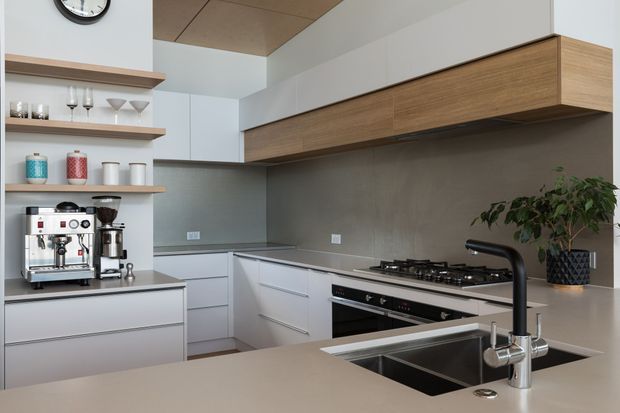 Contemporary L-shaped kitchen with pale grey flat-panel cabinetry, a large island with integrated double sink and tall black tap, stainless-steel appliances, grey splashback, and extensive horizontal timber-grain overhead cabinetry. Open shelving displays glassware and kitchen items, while a potted plant adds greenery. The space has a clean, refined and functional aesthetic with warm timber and neutral finishes.